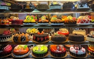 A display case filled with a variety of decorated cakes showcasing intricate designs and vibrant colors. Each cake features unique patterns, toppings, and decorations such as chocolate shavings, fruit slices, and colorful icing. The cakes are arranged neatly in rows across multiple shelves.