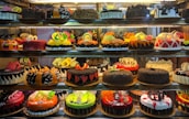 A display case filled with a variety of decorated cakes showcasing intricate designs and vibrant colors. Each cake features unique patterns, toppings, and decorations such as chocolate shavings, fruit slices, and colorful icing. The cakes are arranged neatly in rows across multiple shelves.