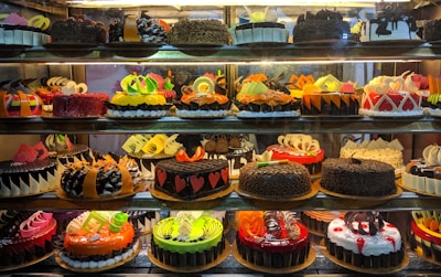 A display case filled with a variety of decorated cakes showcasing intricate designs and vibrant colors. Each cake features unique patterns, toppings, and decorations such as chocolate shavings, fruit slices, and colorful icing. The cakes are arranged neatly in rows across multiple shelves.