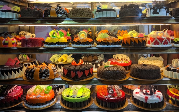 A display case filled with a variety of decorated cakes showcasing intricate designs and vibrant colors. Each cake features unique patterns, toppings, and decorations such as chocolate shavings, fruit slices, and colorful icing. The cakes are arranged neatly in rows across multiple shelves.
