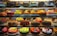 A display case filled with a variety of decorated cakes showcasing intricate designs and vibrant colors. Each cake features unique patterns, toppings, and decorations such as chocolate shavings, fruit slices, and colorful icing. The cakes are arranged neatly in rows across multiple shelves.