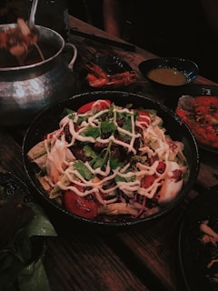 A hearty bowl of salad topped with mixed greens, tomatoes, and drizzles of creamy dressing. Surrounding the main dish are bowls of dipping sauces and what appears to be hot pot ingredients, including prawns and vegetables. The setting is a rustic wooden table, contributing to a warm and inviting atmosphere.