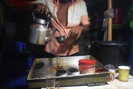 A person is pouring tea from a metal kettle into a glass through a strainer. The setting appears to be a small tea stall or shop, with various containers and cups placed on a wooden counter.