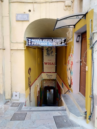 An entrance to a public passage leading to the old port. The corridor is adorned with vibrant murals and signs featuring a Mexican revolution theme. A prominent sign says 'ACCÈS VIEUX PORT PASSAGE PUBLIC.' Below, a narrow stairway extends downward, with another sign referencing the Mexican revolution.