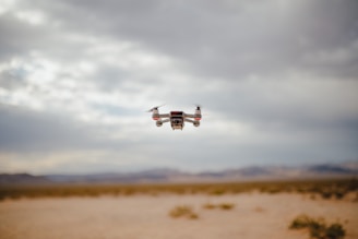 A small drone hovers in the air over a desert landscape. The sky is overcast with clouds, and the ground below is sandy and barren with distant mountains in the background.