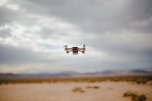 A small drone hovers in the air over a desert landscape. The sky is overcast with clouds, and the ground below is sandy and barren with distant mountains in the background.