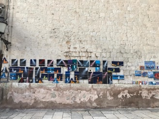 A rustic stone wall serves as the backdrop for an array of colorful artwork displayed in a grid pattern. The artwork includes mostly rectangular and some triangular pieces, featuring a variety of abstract designs and vibrant colors. A metal lamp is mounted on the wall to the left. Below, the ground is paved with light-colored tiles.