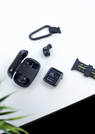 A flat lay photo featuring a black smartwatch with a visible display showing Bluetooth devices, a pair of black wireless earbuds in a glossy case, a single earbud outside, a carabiner clip, and a black silicone watch strap with green accents. The items are arranged on a white surface with a glimpse of a green plant in the corner.
