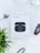A box labeled 'Latitude Series' containing wireless earbuds is positioned in the center on a light background. An additional earbud, a smart watch with colorful display, a set of silver over-ear headphones and a green plant are arranged around it.