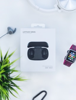 A box labeled 'Latitude Series' containing wireless earbuds is positioned in the center on a light background. An additional earbud, a smart watch with colorful display, a set of silver over-ear headphones and a green plant are arranged around it.