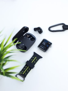 A collection of tech gadgets including wireless earbuds with an open charging case, a smartwatch displaying information, a carabiner clip, and a smartwatch strap. A green plant is partially visible on the side, providing a contrast to the predominantly white background.