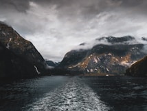 A dramatic landscape with towering, rugged mountains partially shrouded in mist. The foreground features a large body of water, possibly a fjord, with gentle waves indicating movement. The sky is overcast, creating a moody atmosphere as sunlight intermittently breaks through to illuminate patches of the mountain surfaces.