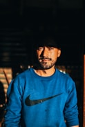 A man with a beard stands looking confidently at the camera, wearing a blue sweatshirt with a recognizable logo. The lighting creates a dramatic effect with shadows emphasizing his facial features.