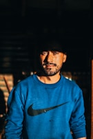 A man with a beard stands looking confidently at the camera, wearing a blue sweatshirt with a recognizable logo. The lighting creates a dramatic effect with shadows emphasizing his facial features.