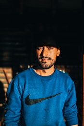 A man with a beard stands looking confidently at the camera, wearing a blue sweatshirt with a recognizable logo. The lighting creates a dramatic effect with shadows emphasizing his facial features.