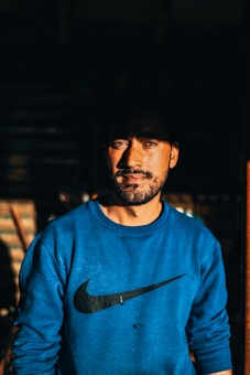 A man with a beard stands looking confidently at the camera, wearing a blue sweatshirt with a recognizable logo. The lighting creates a dramatic effect with shadows emphasizing his facial features.
