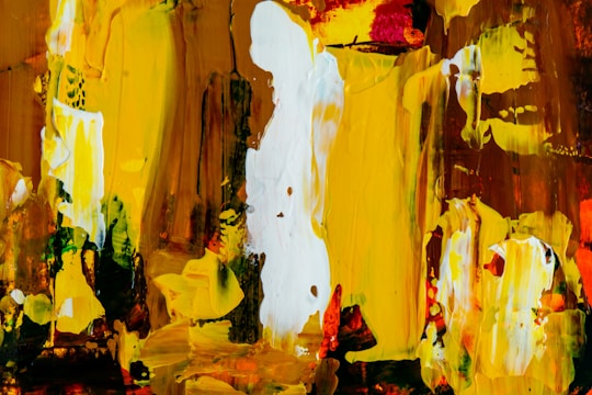 An abstract painting featuring bold strokes and splashes of color. The piece consists predominantly of vibrant yellows, earthy browns, and bright whites with some touches of red and green. The textures are thick and expressive, creating a dynamic and energetic composition.