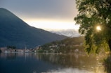A tranquil lake is surrounded by lush green hills and mountains. In the distance, snow-capped peaks are visible under a serene sky. A small, picturesque village lies along the shoreline, reflecting in the calm water. Sunlight peeks through the branches of a large tree, adding a warm glow to the scene.