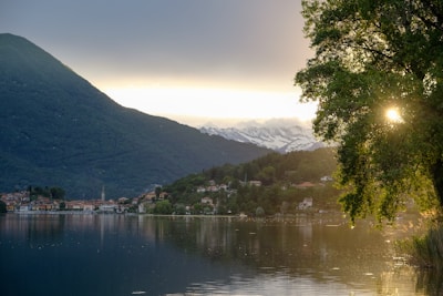 A tranquil lake is surrounded by lush green hills and mountains. In the distance, snow-capped peaks are visible under a serene sky. A small, picturesque village lies along the shoreline, reflecting in the calm water. Sunlight peeks through the branches of a large tree, adding a warm glow to the scene.