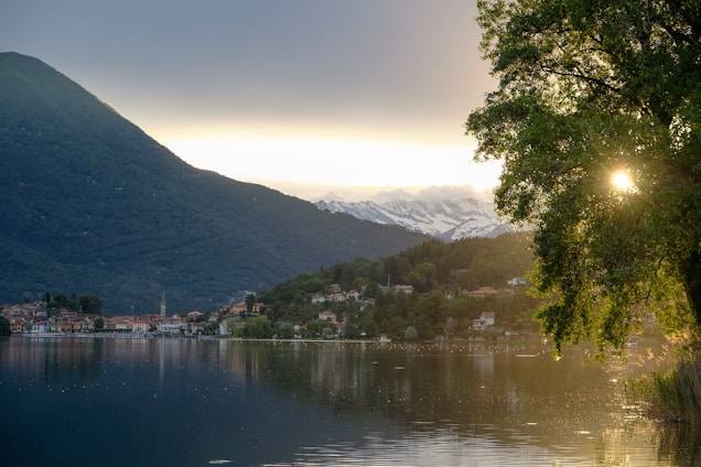 A tranquil lake is surrounded by lush green hills and mountains. In the distance, snow-capped peaks are visible under a serene sky. A small, picturesque village lies along the shoreline, reflecting in the calm water. Sunlight peeks through the branches of a large tree, adding a warm glow to the scene.