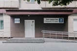 An entrance to a building featuring a light-colored, tiled wall with a ramp and steps leading up to a door. Signs with Cyrillic writing and a logo are present, indicating an organization's name. The area is neatly maintained with a clean courtyard.