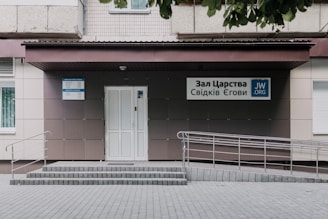 An entrance to a building featuring a light-colored, tiled wall with a ramp and steps leading up to a door. Signs with Cyrillic writing and a logo are present, indicating an organization's name. The area is neatly maintained with a clean courtyard.