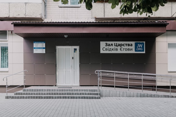 An entrance to a building featuring a light-colored, tiled wall with a ramp and steps leading up to a door. Signs with Cyrillic writing and a logo are present, indicating an organization's name. The area is neatly maintained with a clean courtyard.