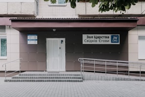 An entrance to a building featuring a light-colored, tiled wall with a ramp and steps leading up to a door. Signs with Cyrillic writing and a logo are present, indicating an organization's name. The area is neatly maintained with a clean courtyard.