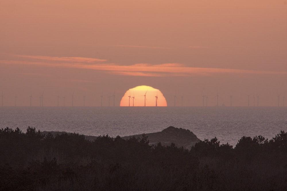 the sun is setting over the ocean with windmills in the distance