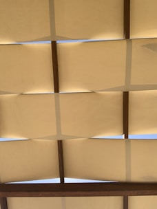 A series of beige fabric panels stretched across a wooden frame with gaps allowing patches of blue sky to be visible between them.
