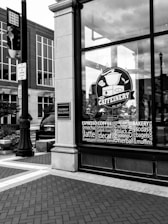 A black and white photograph depicts the storefront of a cafe called 'The Caffeinery' with a large logo featuring a coffee pot. The window is filled with words related to coffee and bakery items. It's located in a brick building, and nearby is a street sign and a lamppost.