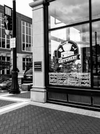 A black and white photograph depicts the storefront of a cafe called 'The Caffeinery' with a large logo featuring a coffee pot. The window is filled with words related to coffee and bakery items. It's located in a brick building, and nearby is a street sign and a lamppost.