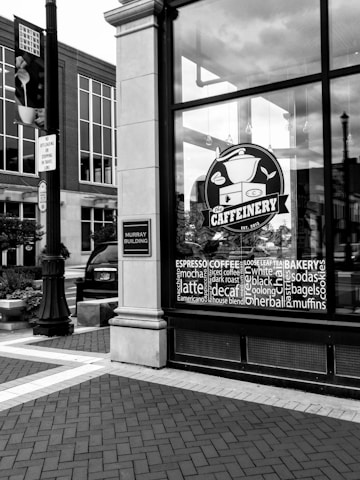 A black and white photograph depicts the storefront of a cafe called 'The Caffeinery' with a large logo featuring a coffee pot. The window is filled with words related to coffee and bakery items. It's located in a brick building, and nearby is a street sign and a lamppost.