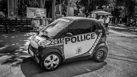 A small, black and white police-themed vehicle is parked on a street. The vehicle features police markings and text in multiple languages, including a badge and the year 2015. People can be seen in the background near a street vendor with an umbrella. Trees and benches line the area, indicating a park or public space.
