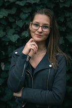 A person with glasses stands confidently in front of a backdrop of dark green ivy leaves. They are wearing a dark jacket with zippers and have a slight smile.