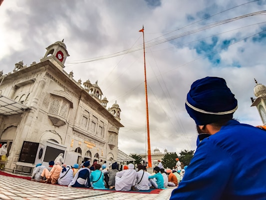 A group of people seated on a checkered floor in front of an ornately designed white building with domes and arches, likely a religious or cultural site. A tall flagpole extends into the cloudy sky. Individuals are wearing colorful turbans and garments, suggesting cultural or religious significance.