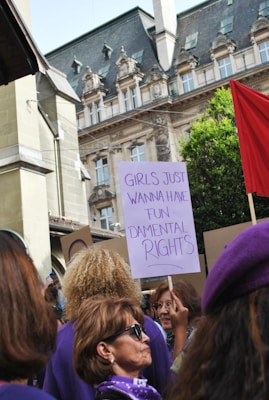 A group of people is gathered outdoors in what appears to be a demonstration or protest. One person is holding a sign that reads 'Girls Just Wanna Have Fundamental Rights.' The background features an ornate building with architectural details and greenery. Most participants appear to be wearing purple items, such as clothing or accessories.