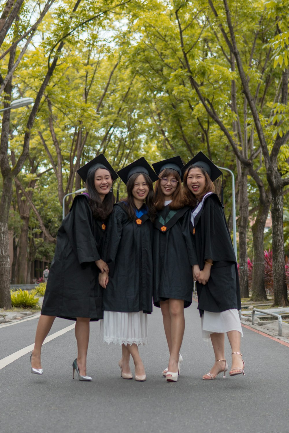 four women in blac academic dresses standing on road between trees