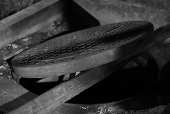 A partially open manhole cover in grayscale, with a textured surface and an underground opening visible. The scene is dimly lit, creating shadows and highlights on the rugged concrete and metal surfaces.