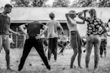 A group of people engaged in a physical activity or exercise routine outdoors. The scene features individuals in various poses and positions, seemingly participating in a coordinated exercise session. The setting appears to be a yard or garden with a backdrop of trees and a wall.