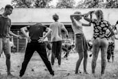 A group of people engaged in a physical activity or exercise routine outdoors. The scene features individuals in various poses and positions, seemingly participating in a coordinated exercise session. The setting appears to be a yard or garden with a backdrop of trees and a wall.