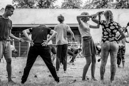 A group of people engaged in a physical activity or exercise routine outdoors. The scene features individuals in various poses and positions, seemingly participating in a coordinated exercise session. The setting appears to be a yard or garden with a backdrop of trees and a wall.