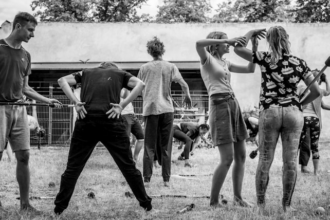 A group of people engaged in a physical activity or exercise routine outdoors. The scene features individuals in various poses and positions, seemingly participating in a coordinated exercise session. The setting appears to be a yard or garden with a backdrop of trees and a wall.