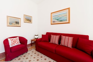A cozy living room corner features a red couch and matching armchair. The couch is adorned with patterned cushions, and a small wooden side table holds a stylish lamp. Three landscape paintings are hung on the white walls, adding a touch of elegance. The floor has a geometric-patterned rug, completing the warm and inviting atmosphere.