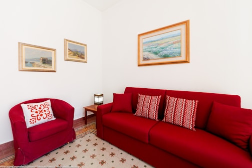A cozy living room corner features a red couch and matching armchair. The couch is adorned with patterned cushions, and a small wooden side table holds a stylish lamp. Three landscape paintings are hung on the white walls, adding a touch of elegance. The floor has a geometric-patterned rug, completing the warm and inviting atmosphere.