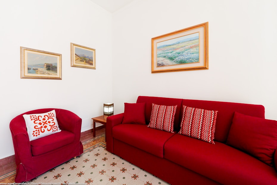 A cozy living room corner features a red couch and matching armchair. The couch is adorned with patterned cushions, and a small wooden side table holds a stylish lamp. Three landscape paintings are hung on the white walls, adding a touch of elegance. The floor has a geometric-patterned rug, completing the warm and inviting atmosphere.