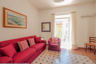 A cozy living room with a red sofa adorned with patterned cushions, and a matching red armchair with a decorative pillow. There is a piece of artwork hanging above the sofa and a mirror on the adjacent wall. The room is bright, benefitting from natural light coming through a window covered with sheer curtains. An air conditioning unit is mounted on the wall above a wooden chair. The floor features decorative tiles.