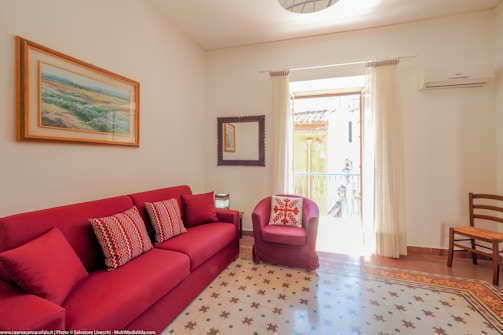 A cozy living room with a red sofa adorned with patterned cushions, and a matching red armchair with a decorative pillow. There is a piece of artwork hanging above the sofa and a mirror on the adjacent wall. The room is bright, benefitting from natural light coming through a window covered with sheer curtains. An air conditioning unit is mounted on the wall above a wooden chair. The floor features decorative tiles.