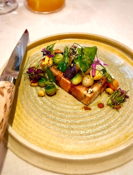 A beautifully arranged gourmet dish sits on a textured ceramic plate. The dish features a colorful assortment of vegetables, vibrant greens, and delicate edible flowers. A polished knife with a decorative handle is placed next to the plate, suggesting an emphasis on presentation and fine dining.