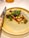 A beautifully arranged gourmet dish sits on a textured ceramic plate. The dish features a colorful assortment of vegetables, vibrant greens, and delicate edible flowers. A polished knife with a decorative handle is placed next to the plate, suggesting an emphasis on presentation and fine dining.